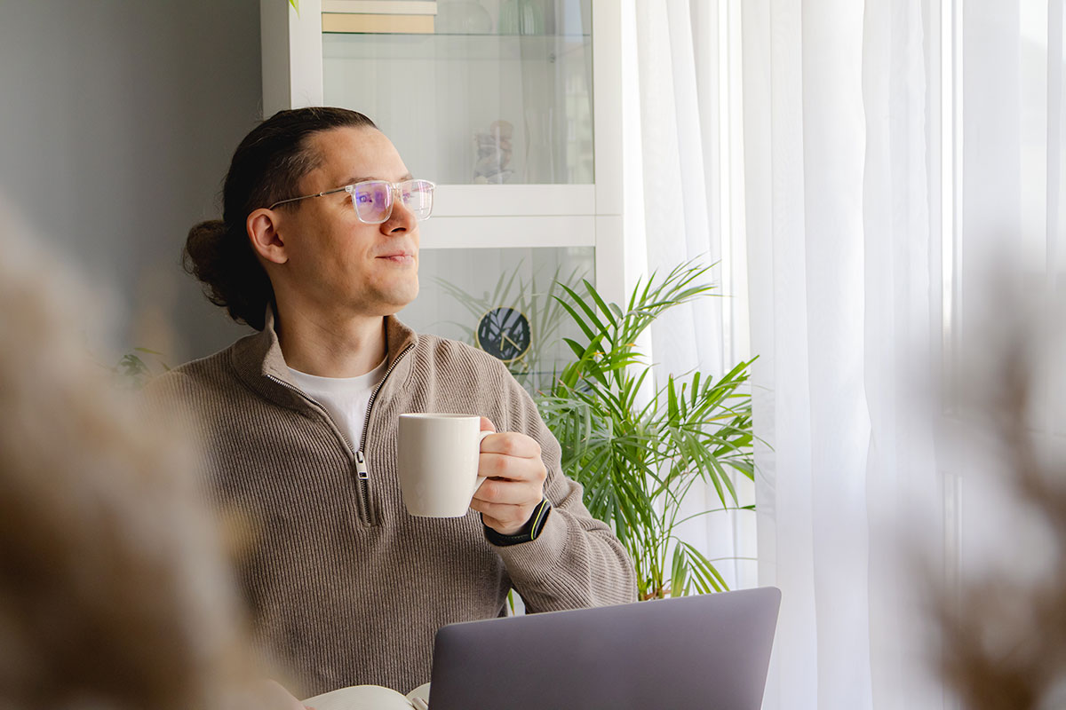 man holding cup of coffee relaxing by window working from home